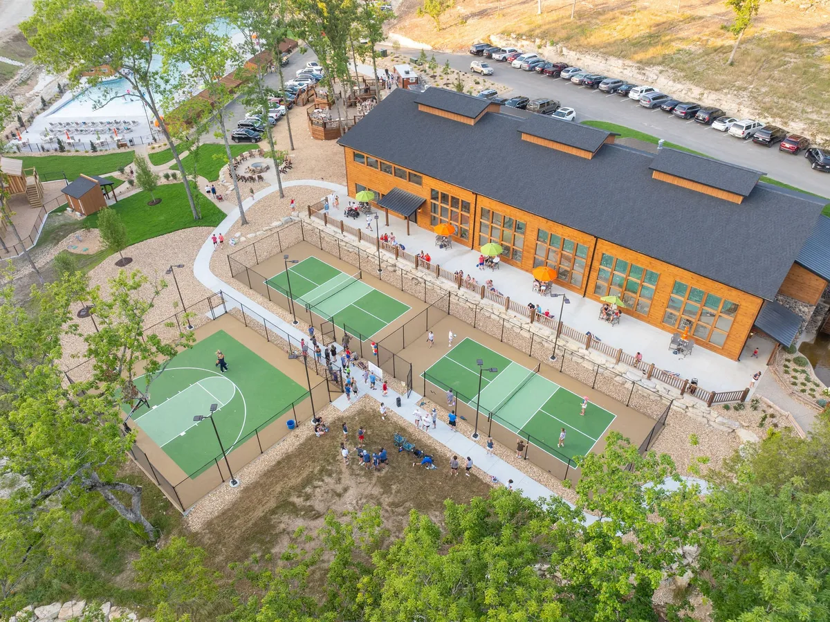 An energetic exterior image showing a large wooden building, adjacent sports courts, and a gathering of people enjoying various activities. The scene is bustling with outdoor basketball and tennis games, as well as groups socializing at tables under the trees.
