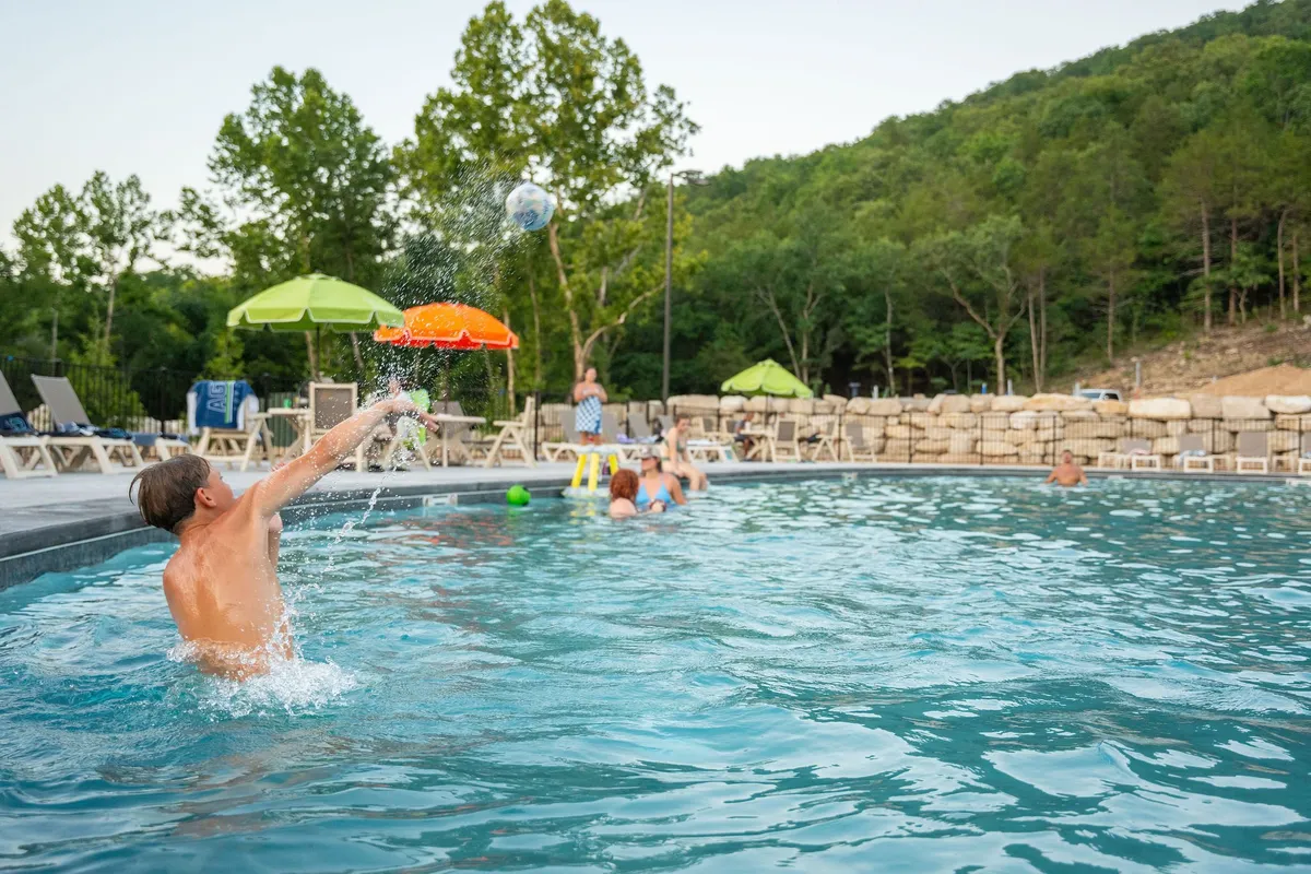 Children are splashing and playing in a community outdoor swimming pool, surrounded by green umbrellas and a rocky landscape. Adults relax nearby, emphasizing a safe and joyful family environment.