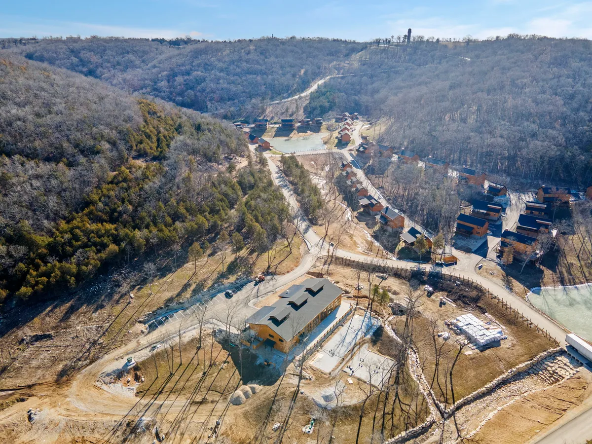 A central lodge is surrounded by newly built cabins, set against a backdrop of trees and open sky. The area exudes a sense of community with wide paths, and the natural setting is inviting. Some minor landscaping and construction touches are still visible, reflecting an area in active development.