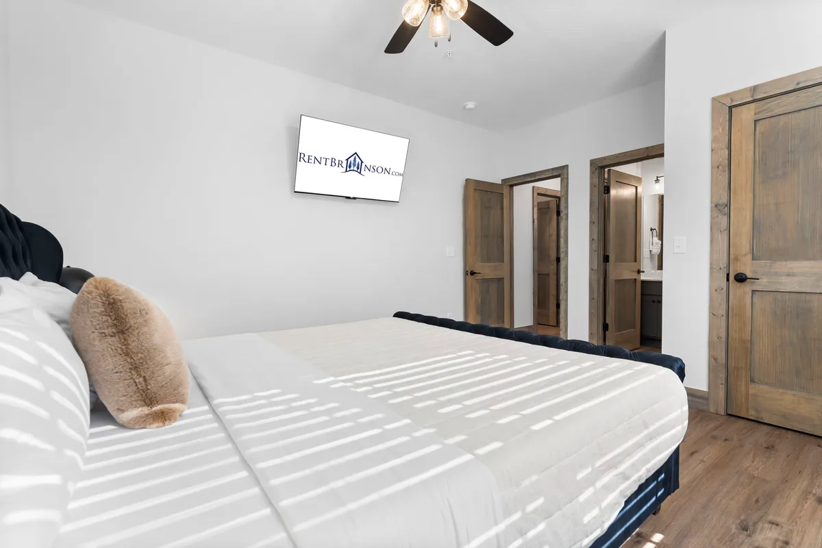 This bedroom shows a bed with crisp white linens, a wall-mounted TV, and wooden doors, projected in a calm, neutral palette. The space is tranquil and uncluttered, enhancing the sense of peaceful retreat.