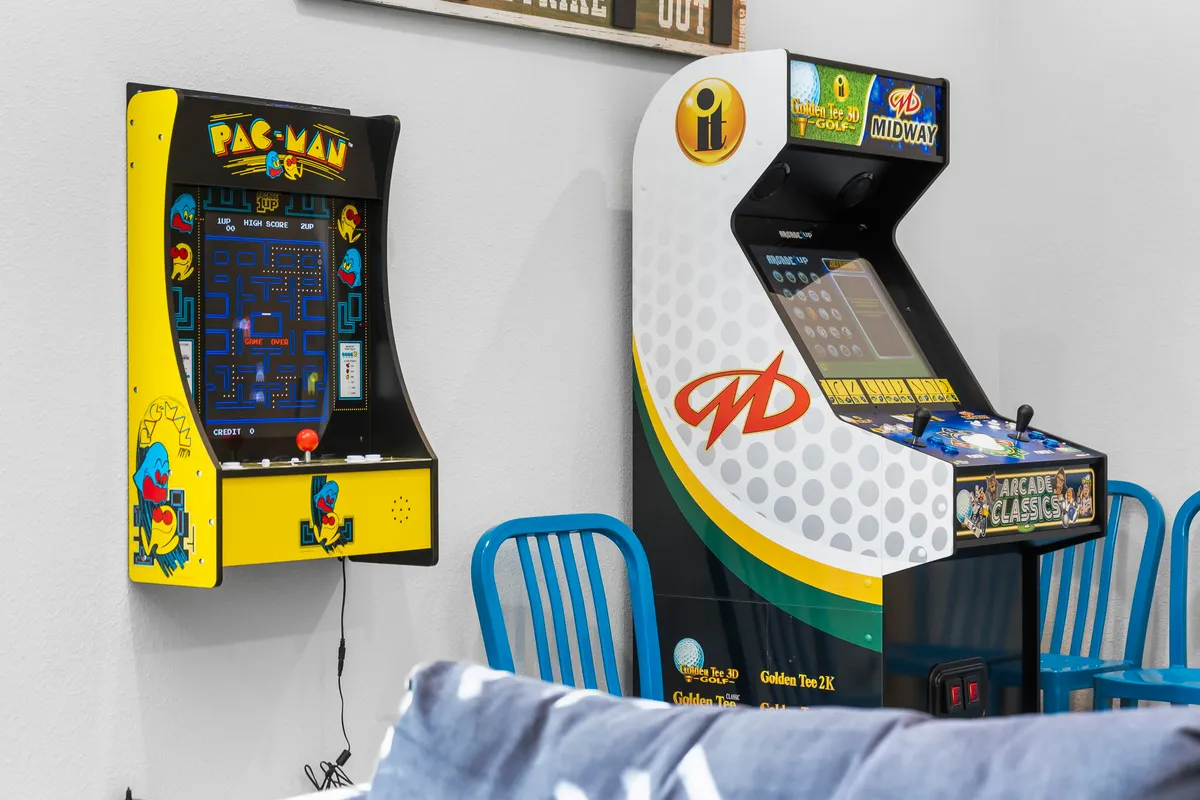A close-up view of a classic Pac-Man arcade machine alongside another Midway arcade cabinet, with blue chairs nearby. The nostalgic setup invites visitors to challenge each other to arcade classics, echoing the section's emphasis on retro gaming and high-score duels.