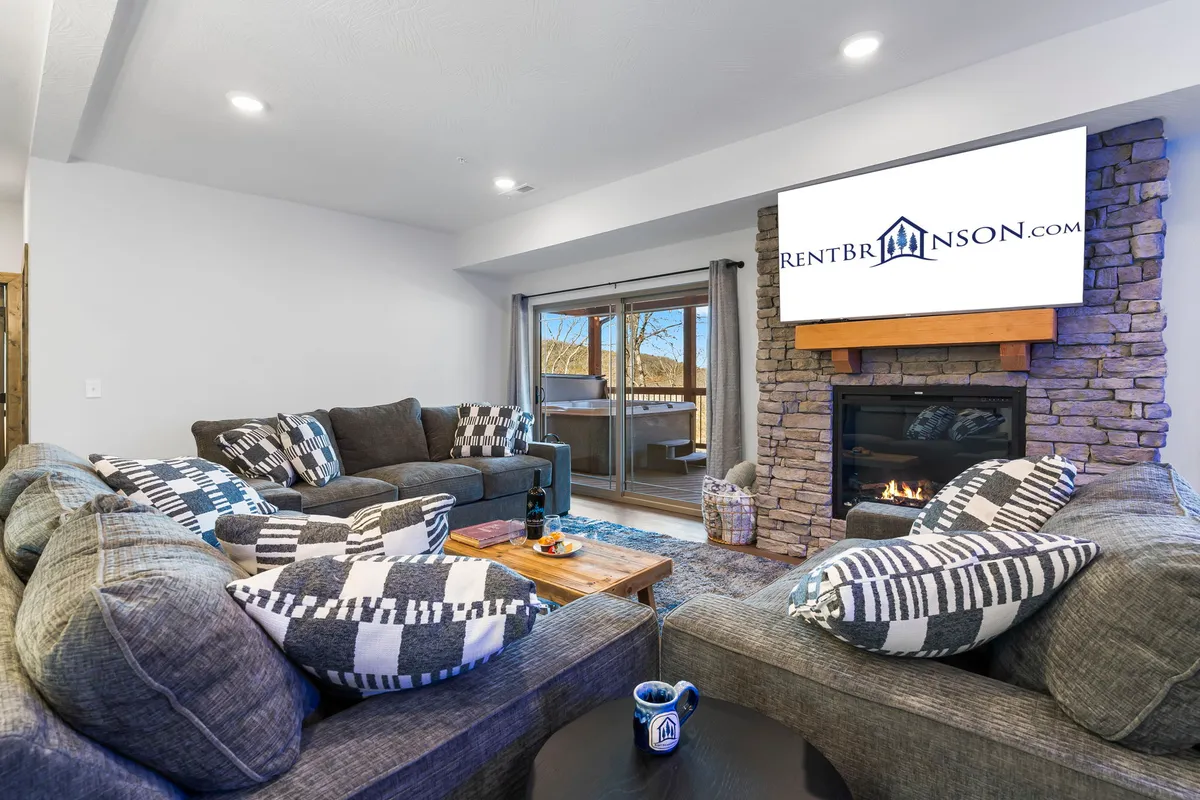 The image showcases the cabin's spacious living area, featuring a plush sectional sofa arranged around a cozy stone fireplace and a large television. A rustic wooden coffee table anchors the room, creating a warm, communal space perfect for relaxation and gathering. The overall ambiance is inviting, with soft lighting and natural wood accents enhancing the comfort and charm.