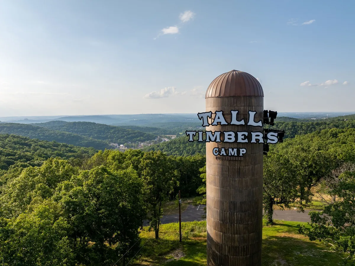 A welcoming sign reading 'Tall Timbers Camp' stands at the entrance to a picturesque setting of rolling lawns and light woods. The scene feels adventurous and fresh, suggesting new beginnings and a gateway to exploration within the community.