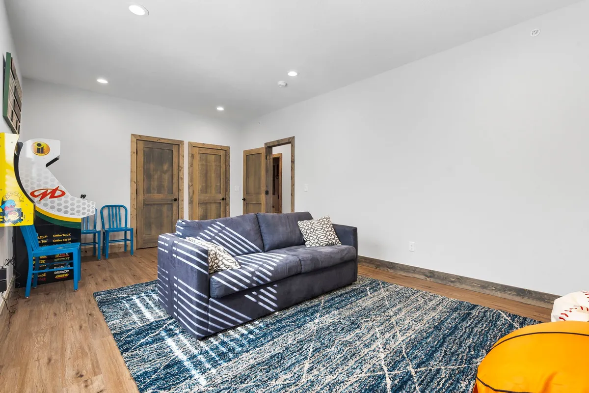 A cozy game lounge featuring a plush sofa, an arcade machine, a patterned rug, and blue accent chairs. The room feels playful yet relaxed, with plenty of space for family or friends to gather and compete in games or unwind together.