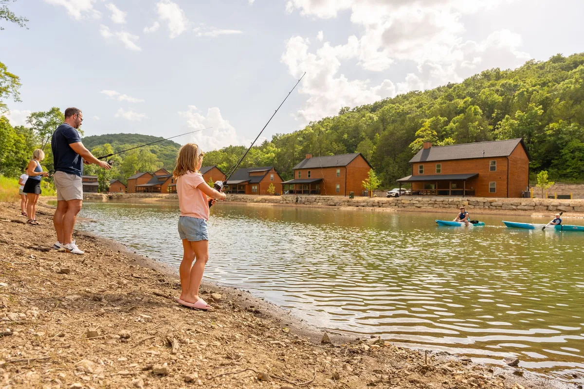 This image shows several people fishing from the bank of a lake and others kayaking on the water, with welcoming cabins visible in the background. The scene is lively yet relaxed, capturing a multigenerational group enjoying outdoor activities together.