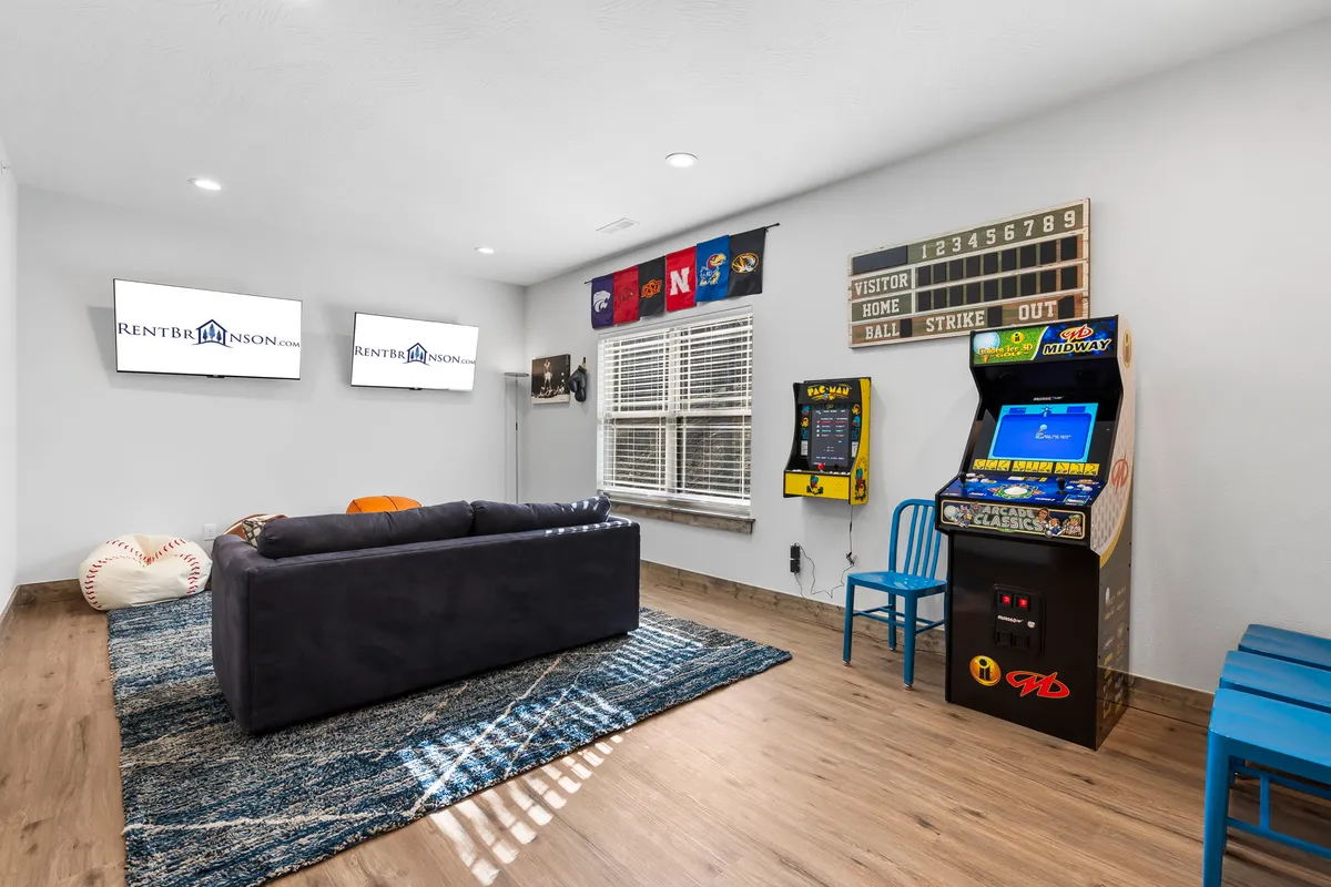A lively game room with a sofa set up for socializing, an arcade game visible in the corner, sports-themed wall decor, and multiple televisions mounted on the wall. The arrangement creates an inviting space for gaming, watching sports, and hanging out with friends or family.