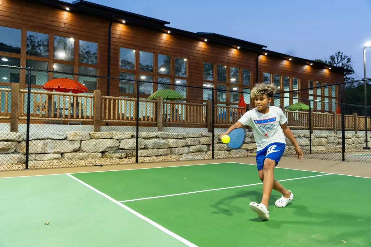 A child is actively playing pickleball on a court with colorful umbrellas and a wooden building in the background, suggesting a vibrant and family-friendly recreational area.